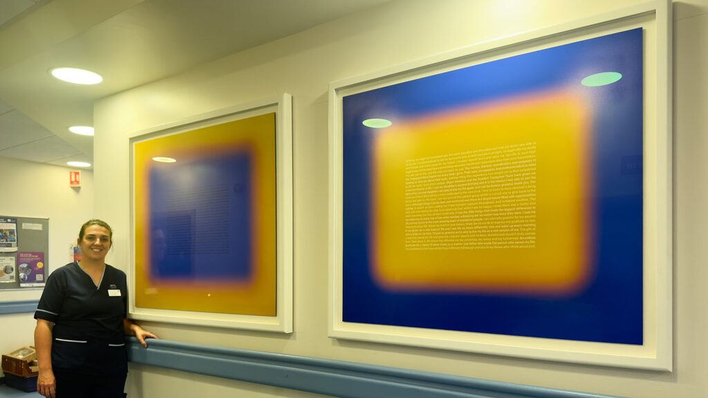 Senior Charge Nurse, Sam Slight, standing beside Grateful by Rebecca Milling in the corridor of the Royal Infirmary of Edinburgh outside the Transplant Unit